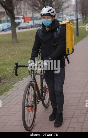 Vertikale Aufnahme eines männlichen Kuriers mit medizinischer Gesichtsmaske und Thermo-Delivery-Rucksack, zu Fuß mit Fahrrad Stockfoto