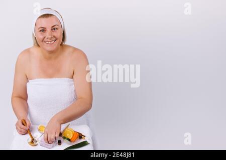 Eine Frau zu Hause, eingewickelt in ein weißes Handtuch mit einem Lächeln, bereitet eine Gesicht und Körpermaske aus natürlichen Produkten. Weißer Hintergrund und leerer Seitenraum. Stockfoto