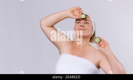 Banner. Ältere Frau mit gepflegter Haut und einem Verband auf ihrem Kopf schaut nach oben und hält Gurkenscheiben in ihren Händen. Home Hautpflege auf einem weißen Stockfoto