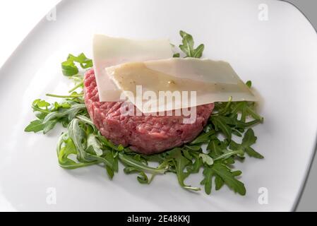 Frisches Rindfleisch Tartare, rohes Hackfleisch aus piemontesischen Fassona Rasse Rinder.mit Käse Flocken, auf Rucola Blätter in weißem Teller Stockfoto