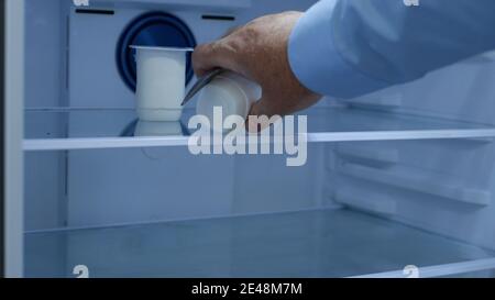 Hungry man öffnet die Kühlschranktür und sucht nach Essen im Inneren, nimmt einen frischen und kalten Joghurt und einen Teelöffel Stockfoto