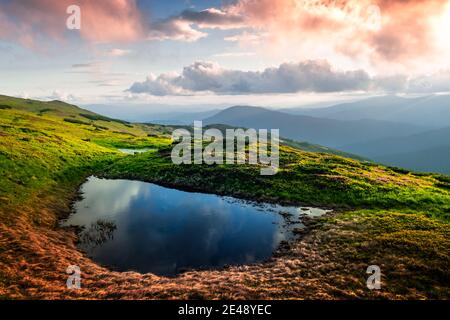 Bergsee bei Sonnenaufgang. Malerische Sommerlandschaft mit grünen Hügeln und Sonnenstrahlen am Morgenhimmel. Karpaten Stockfoto