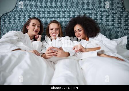 Drei beste Freundinnen chillen im Bett und Scrollen auf dem Smartphone Stockfoto