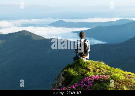 Ein Tourist sitzt am Rande einer Klippe mit einem rosa Teppich von Rhododendron Blumen im Sommer bedeckt. Neblige Berge im Hintergrund. Landschaftsfotografie Stockfoto