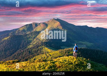 Ein Tourist am Rande eines Berges mit einem üppigen Gras bedeckt. Lila Sonnenuntergang Himmel und Höhe Berge Gipfel auf Hintergrund. Landschaftsfotografie Stockfoto