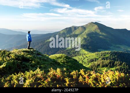 Ein Tourist am Rande eines Berges mit einem üppigen Gras bedeckt. Wolkiger blauer Himmel und hohe Berge Gipfel auf dem Hintergrund. Landschaftsfotografie Stockfoto