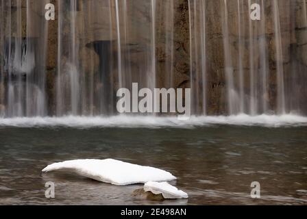 Verschwommenes Wasser, das aus einer Kaskade in einem Bach fällt. Mit Eis im Vordergrund. Stockfoto