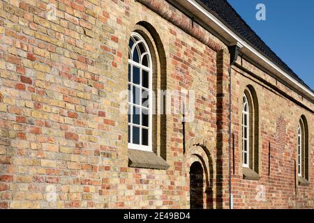 Seitenfassade einer alten restaurierten Kirche mit gewölbten Fenstern und einer Tür in einem Mauerwerk aus alten gelben, orangen und roten Ziegeln vor einem blauen Himmel in Exmorra Stockfoto