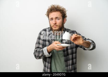 Gut aussehender Mann mit lockigem Haar und Bart halten umgekehrte Tasse zeigt keinen Kaffee, Kerl trägt karierte Langarm-Shirt isoliert auf weißem Hintergrund