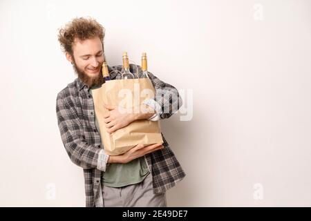 Junger Mann, der eine Einkaufstasche aus Papier mit lachenden Weinflaschen trägt und darauf hinunterblickt. Hübscher junger Mann mit lockigen Haaren in olivem T-Shirt Stockfoto