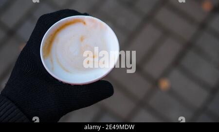Ein Glas Cappuccino in der Hand eines mannes trinkt ein junger Mann Cappuccino, während er in Winterhandschuhen eine Tasse Kaffee hält. Stockfoto