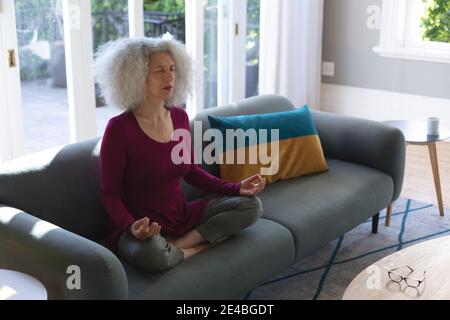Ältere kaukasische Frau sitzt auf der Couch im Wohnzimmer meditieren. Zu Hause bleiben in Selbstisolation während Quarantänesperre. Stockfoto