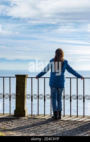 Frau in einer blauen Daunenjacke auf dem Geländer vorbei Ein See Stockfoto