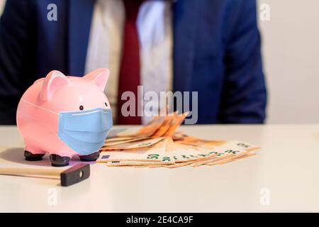 Nahaufnahme von Sparschwein, Tragen einer schützenden Gesichtsmaske. Sparkonzept zur Zeit der Coronavirus-Pandemie. Fünfzig Euro und Geschäftsmann im Hintergrund Stockfoto