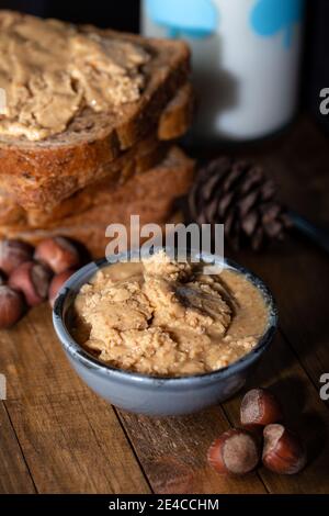 Hausgemachte Nussbutter in einer kleinen Schüssel. Brotscheiben mit hausgemachtem Haselnussaufstrich und einem Glas Milch auf einem Holztisch. Hausgemachte Haselnussbutter. Stockfoto