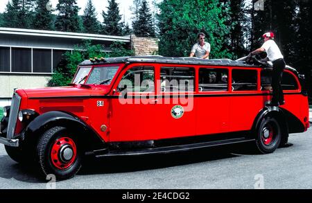 Zwei Männer in diesem langen roten Reisebus aus den 1930er Jahren öffnen sein Rollback-Segeltuch-Dach, um Touristen, die malerische Ausflüge im Glacier National Park im Nordwesten von Montana, USA, Unternehmen, einen Panoramablick auf beeindruckende Berggipfel zu bieten. Das 17-Personen-Fahrzeug gehört zu den 33 identischen Bussen des Parks, die als Red Jammers bekannt sind, weil ihre Fahrer beim Doppelschalten der Schaltgetriebe auf den steilen Bergstraßen lautstark die Gänge „verklemmen“. Alle historischen Modelle der White Motor Company No. 706 wurden in ihr ursprüngliches Aussehen zurückversetzt und mit Automatikgetrieben nachgerüstet. Stockfoto