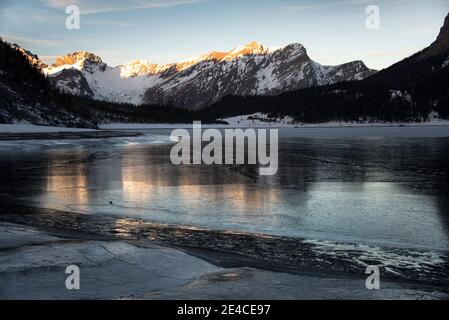 Gefrorener Bergsee im Morgengrauen erleuchten die ersten Sonnenstrahlen die Gipfel Stockfoto