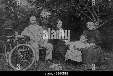 Amerikanisches Archiv monochromes Familienportrait in einem Garten mit Generationen von Urgroßmutter, Großeltern, Mutter und Tochter mit Hund und Fahrrad, aufgenommen im späten 19. Jahrhundert in Port Byron, NY, USA Stockfoto