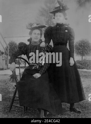 Amerikanisches Archiv monochromes Porträt zweier Frauen in passenden Tartan-Outfits in einem Garten. Eine junge Frau steht und eine Frau sitzt auf einem Stuhl. Aufgenommen im späten 19. Jahrhundert in Port Byron, NY, USA Stockfoto