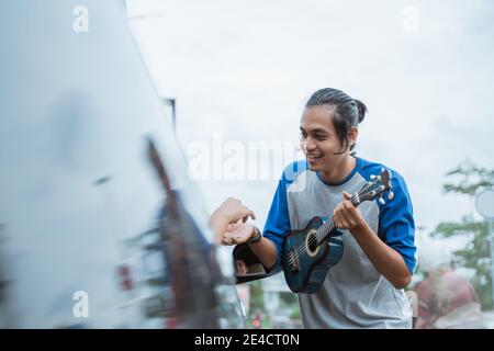 Der Busker, der die Ukulele hält, wenn er eine Münze von erhält Jemand gibt aus dem Auto auf der Seite des Straße Stockfoto