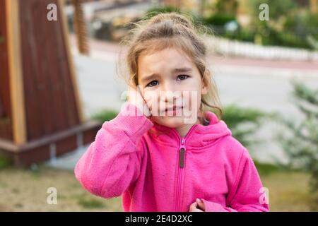 Ein kleines Mädchen in einem rosa Hoodie mit einem traurigen und tränenreichen Gesicht hält ihr Ohr. Ohrschmerzen, Otitis media, Schwellung der Wange, Zahnfleisch, Zahnschmerzen, Kinderhaut Stockfoto