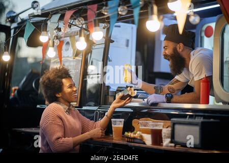 Junge bärtige kaukasische Angestellte in Fast Food lachend, Zugabe eines Senf in einem Sandwich zu einer weiblichen afro-amerikanischen zufriedenen Kunden Stockfoto