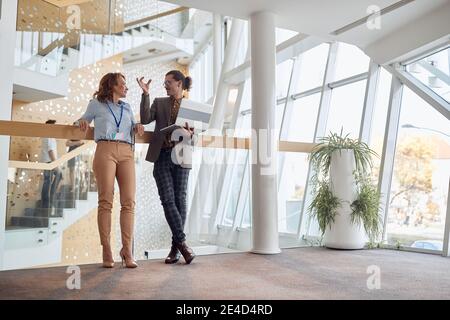 Junger Mann erklärt älteren weiblichen Kollegen im Flur eines Geschäftsgebäudes stehend, lehnte sich an einen Zaun, Stockfoto