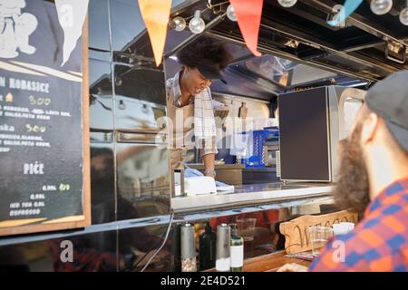Junge afro-amerikanische Mitarbeiterin in Fast-Food-Service schreiben Der Auftrag von einem jungen kaukasischen bärtigen Hipster mit einem Kap Stockfoto