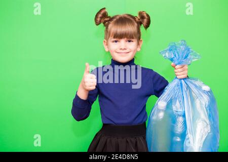 Happy kleines Mädchen hält Müllbeutel mit Plastikmüll und Zeigt die Geste des Gewinnens isoliert auf grünem Hintergrund Stockfoto