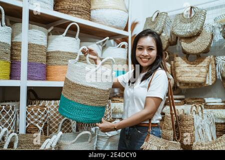 Frau lächelt an der Kamera, während sie ein Gewebe hebt Korb steht unter den Bastelgegenständen in der Handwerkergalerie Stockfoto