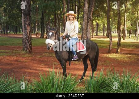 Junge Frau in Hemd und Strohhut, Reiten dunkelbraun Pferd im Park, verschwommen hohen Kiefern im Hintergrund Stockfoto