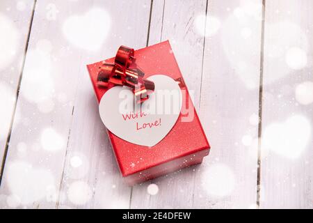 Rote Box mit einer großen herzförmigen Karte für ein besonderes Valentinsgeschenk. Weißer Holzhintergrund und Bokeh-Effekt rundum. Stockfoto