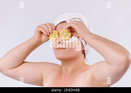 Porträt einer älteren Frau mit einem Handtuch auf dem Kopf, schaut zur Seite und hält Zitronenscheiben vor ihren Augen. Weißer Hintergrund. Stockfoto