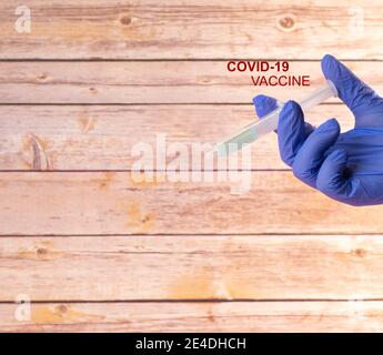 Hand mit einem Krankenhaus Latexhandschuh durch die Einnahme einer Spritze mit dem covid-19 Impfstoff mit einem hölzernen Hintergrund und einem Titel, der covid-19 Impfstoff sagt. Stockfoto