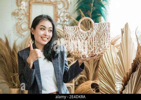 Eine Geschäftsfrau in einem Blazer trägt eine gewebte Tasche mit Daumen nach oben, während vor einem Hintergrund von vielen Handarbeiten stehen Stockfoto