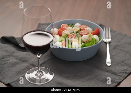 Salat mit Romaine, Tomaten und Mozzarella in blauer Schüssel mit einem Glas Rotwein, flacher Fokus Stockfoto