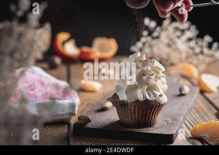 Eine Hand streut Kakaopulver auf einem Cupcake mit Schlagsahne auf einem Holztisch. Stockfoto