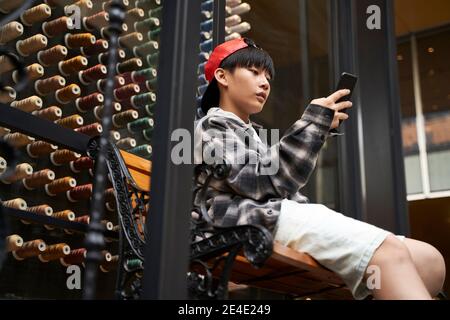 15-jähriger asiatischer Teenager-Junge, der auf der Bank sitzt und Handy anschaut Stockfoto