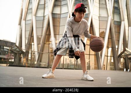 15-jähriger asiatischer Teenager-Junge spielt Basketball im Freien vor Ein modernes Gebäude Stockfoto