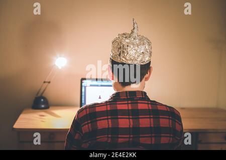 Der junge Mann mit Aluminiumkappe sitzt im dunklen Keller vor einem Laptop. Verschwörungstheorie Konzept Stockfoto