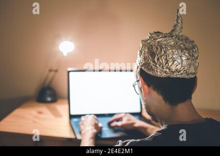 Der junge Mann mit Aluminiumkappe sitzt im dunklen Keller vor einem Laptop. Verschwörungstheorie Konzept Stockfoto