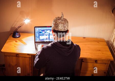 Der junge Mann mit Aluminiumkappe sitzt im dunklen Keller vor einem Laptop. Verschwörungstheorie Konzept Stockfoto