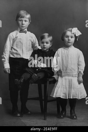 Amerikanisches Archiv monochromes Familienportrait von drei kleinen Kindern: Zwei Jungen und ein Mädchen. Der Junge sitzt auf einem Stuhl und hält ein Spielzeug. Aufgenommen im späten 19. Jahrhundert Stockfoto