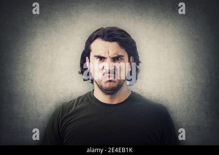 Verwirrt und verwirrt junger Mann, der perplex und aufgeregt auf Kamera isoliert auf grauem Wandhintergrund aussagt. Angry Casual guy, lange lockige Frisur, irr Stockfoto