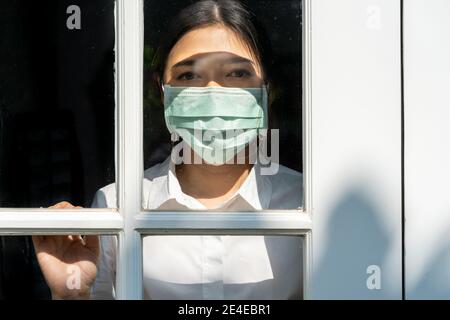Junge Frau trägt chirurgische Maske Aufenthalt in ihrem Haus Blick aus der Haustür in Sorge während COVID Pandamic Sperre, Gesundheitskonzept Stockfoto