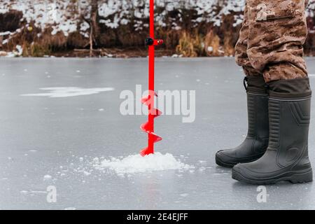 Gesichtsloser Fischer bohrt ein Loch mit einer Eisschraube für das Eisfischen. Angeln im Winter. Mannbeine in Stiefeln und Eisschraube auf Eishintergrund Stockfoto