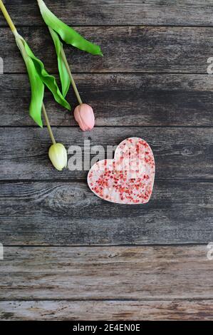 Eine herzförmige rosa Schokolade mit getrockneten Erdbeeren auf einem Vintage dunklen Holztisch Hintergrund mit Tulpenblüten. Liebe und Grußkarte Konzept. Stockfoto