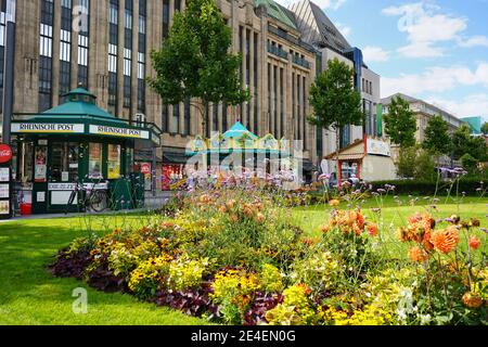 Blick auf den Corneliusplatz in der Düsseldorfer Innenstadt mit Zeitungskioskade und nostalgischem Karussell vor dem Kaufhof-Gebäude. Stockfoto