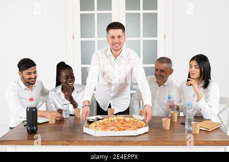 Das Business Team isst Pizza im Büro und sitzt um den Tisch. Überglücklich gemischte Mitarbeiter der Rennfirma versammelten sich am Bürotisch Stockfoto