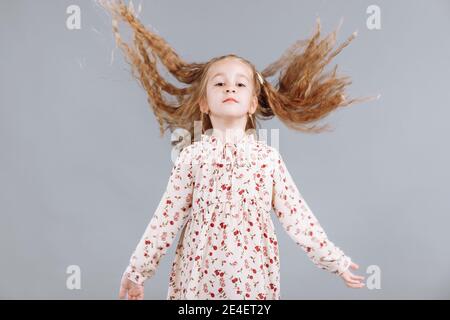 Porträt eines schönen lockigen kleinen Mädchens in einem eleganten Blumenkleid steht, lacht und schaut in die Kamera im Studio auf einem grauen Hintergrund Stockfoto